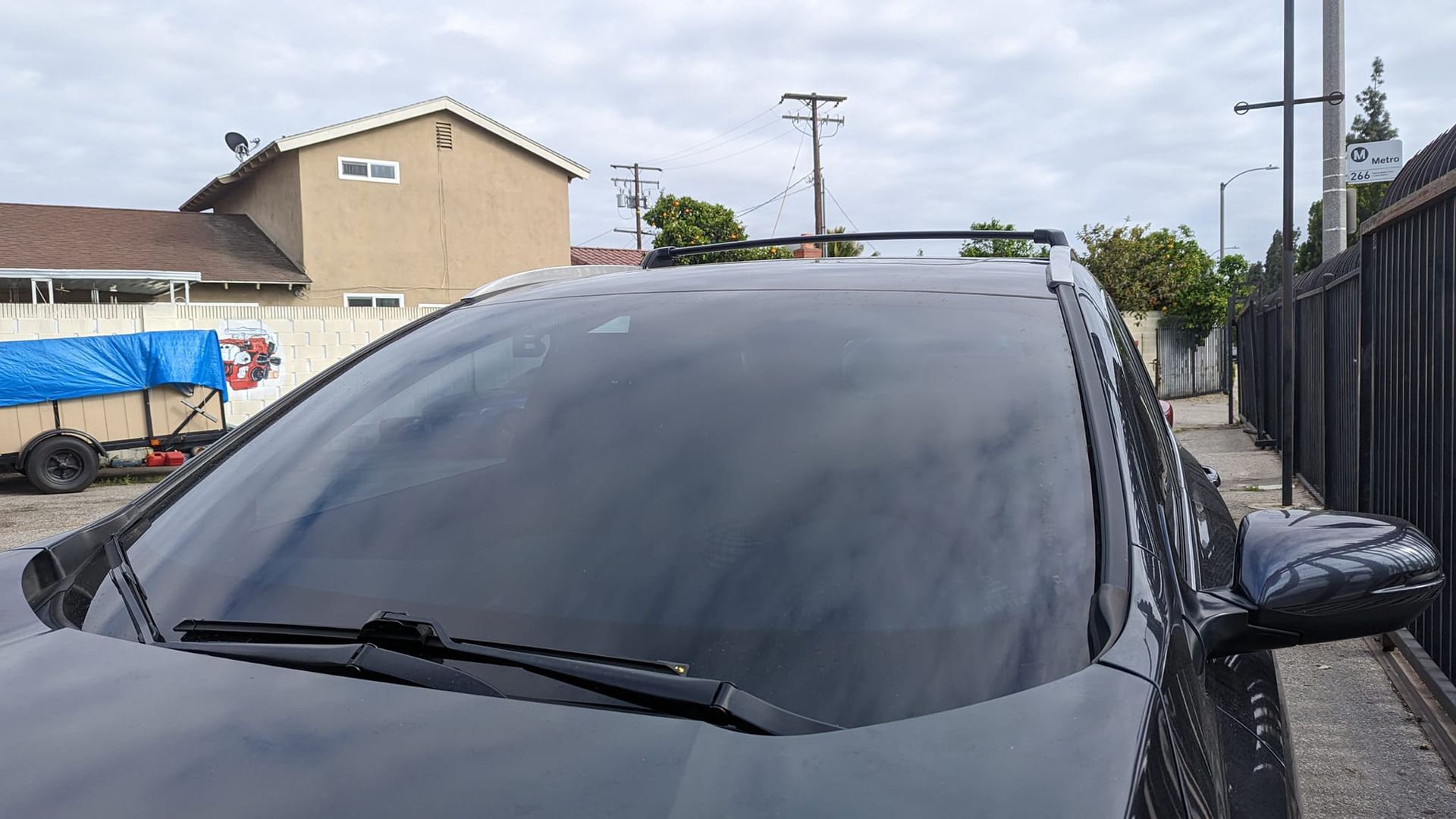 Black car with roof rack parked outdoors; gray sky and buildings in background.