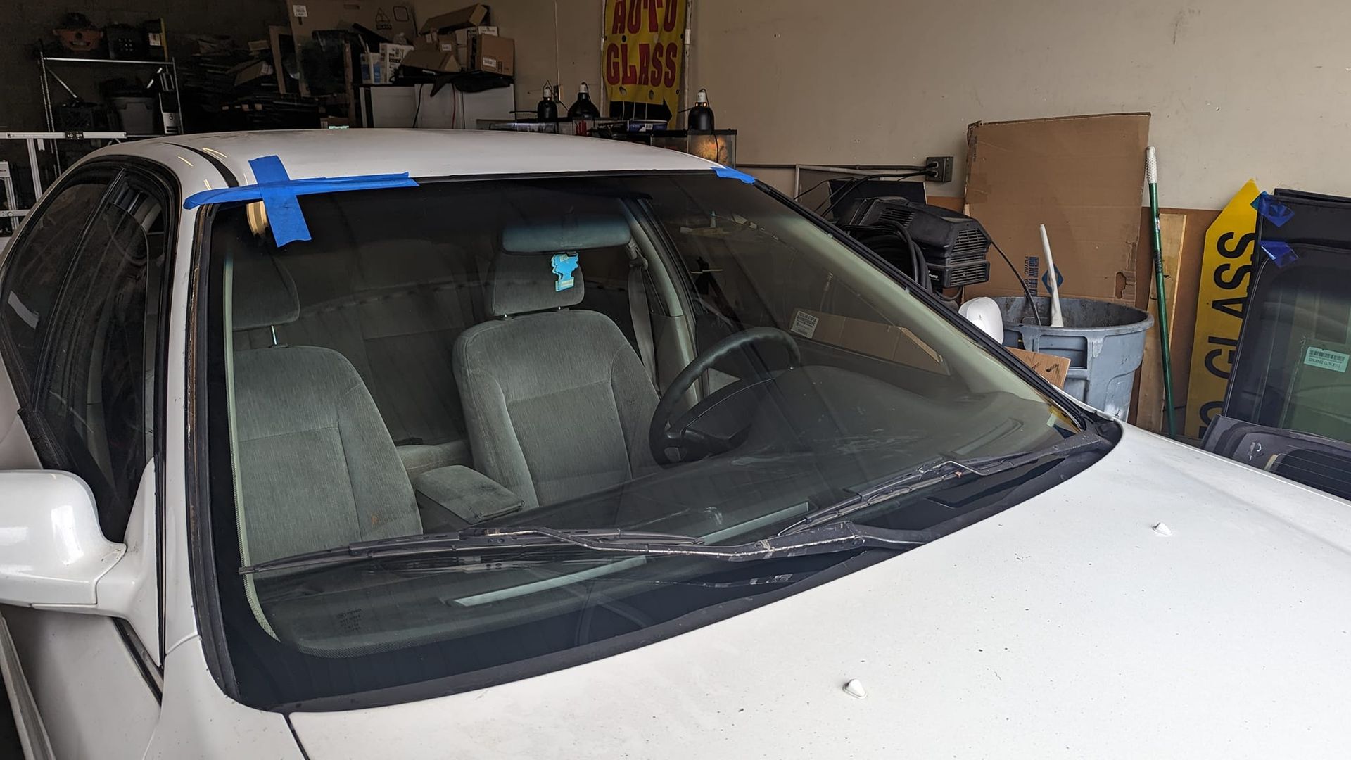 White car with blue tape on windshield, parked in a garage.