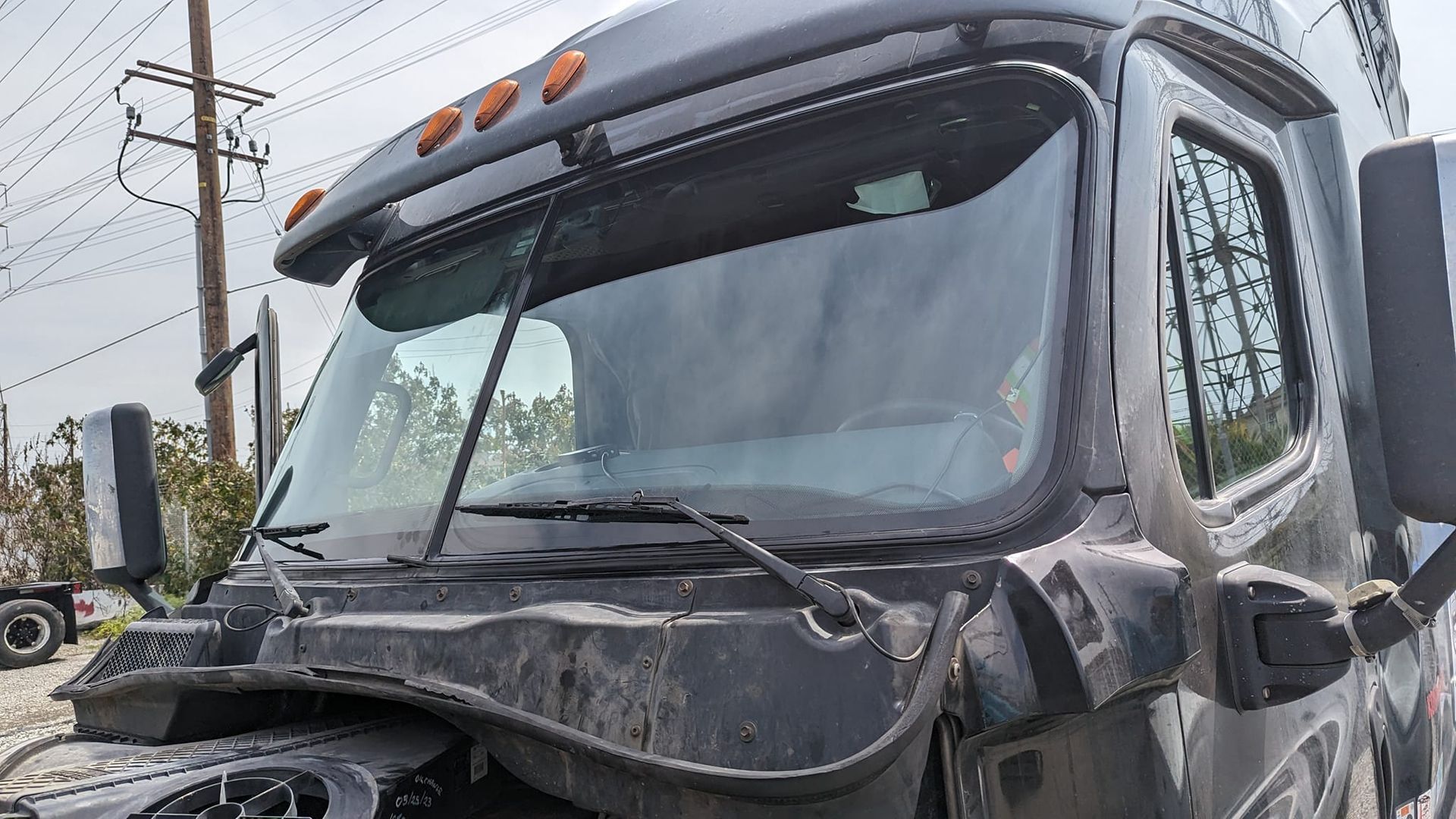Close-up of a damaged dark-colored semi-truck cab, windshield and surrounding area. Debris and damage are visible.