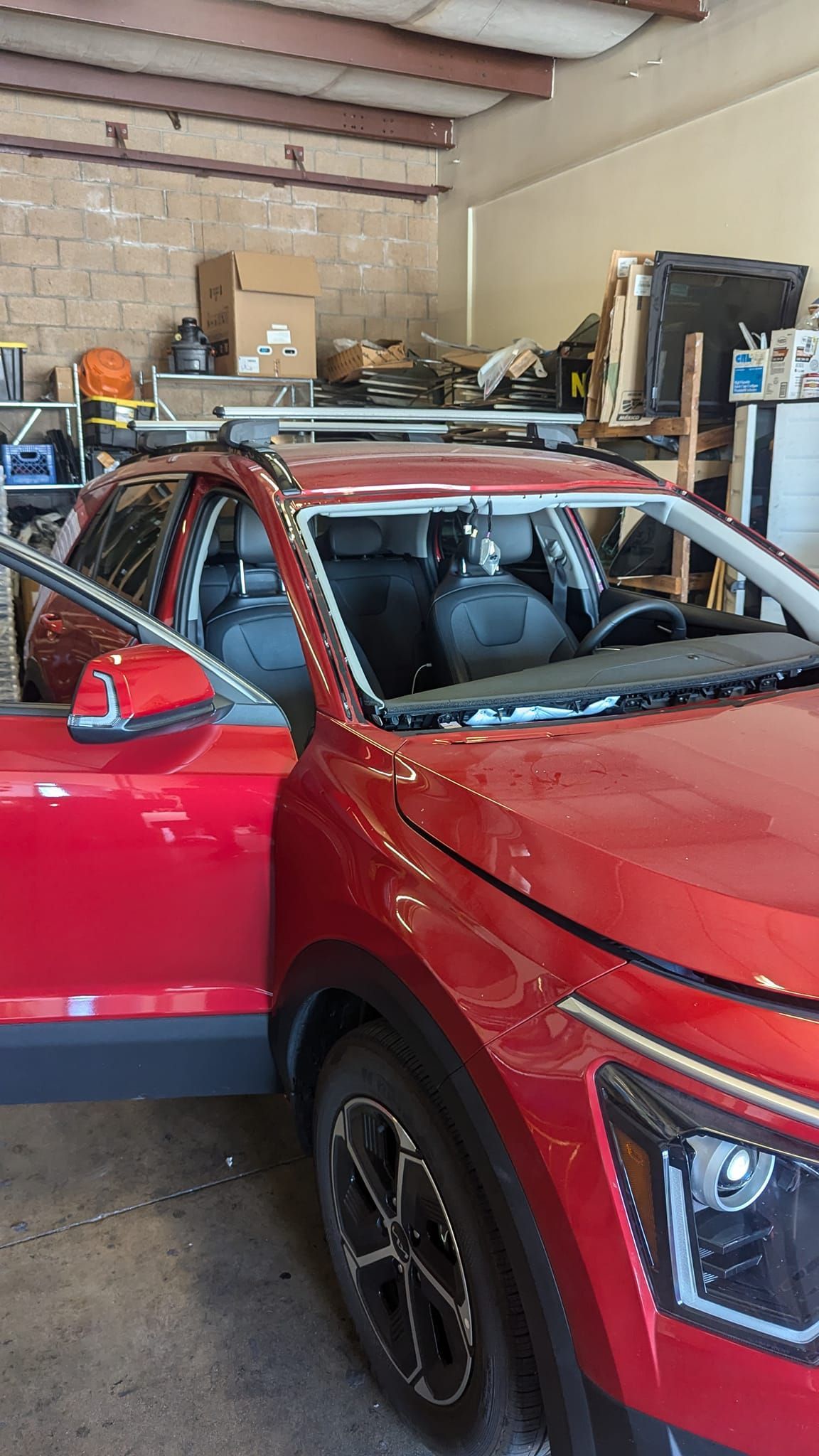 Red car with open door in a garage; broken windshield, roof rack installed.