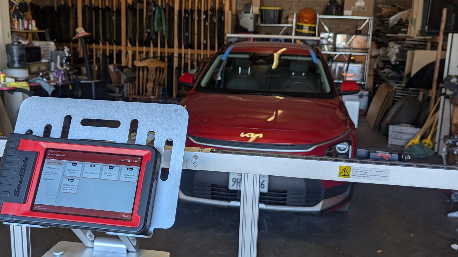 Red car undergoing wheel alignment in a garage. Alignment machine in front; tools and equipment visible.