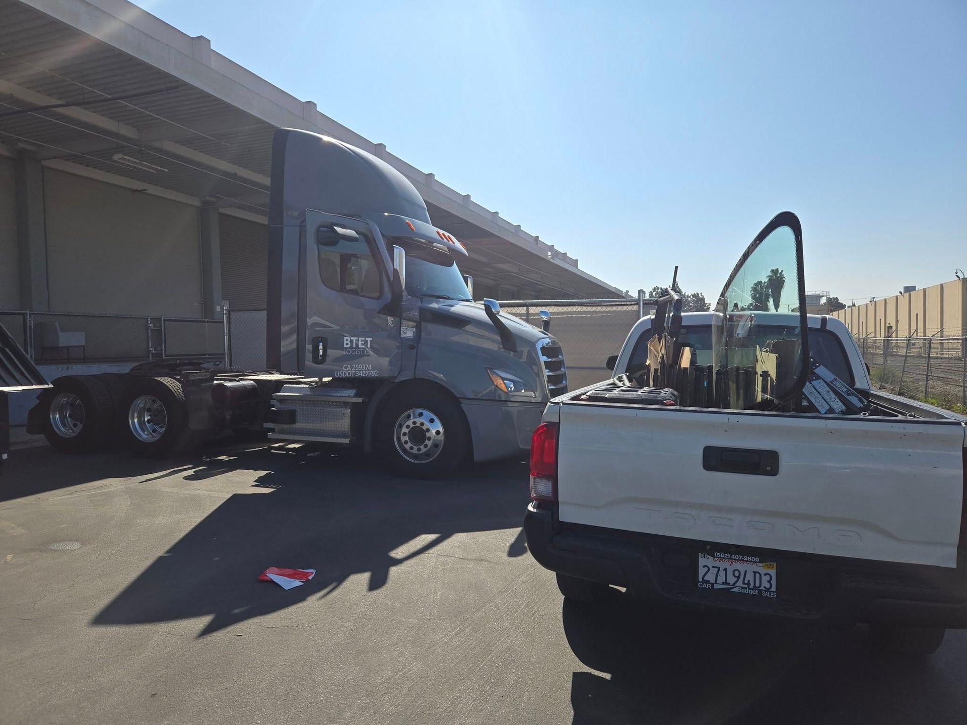 Grey semi-truck next to a white pickup truck. Both parked near a loading dock in sunlight.