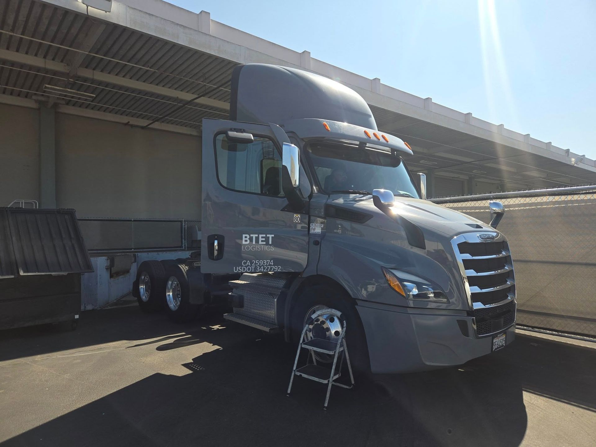 Gray semi-truck with open door parked under a covered area, sunny day.