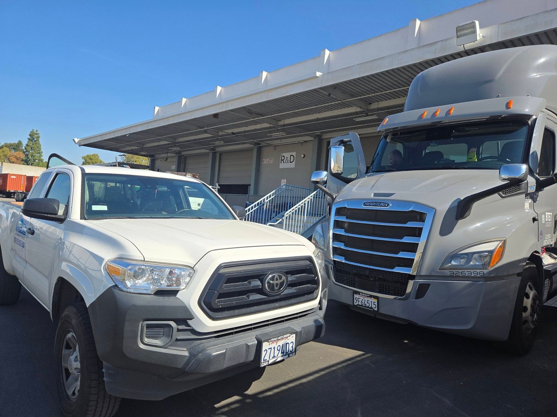 White pickup truck and gray semi-truck parked in front of loading docks under a bright blue sky.