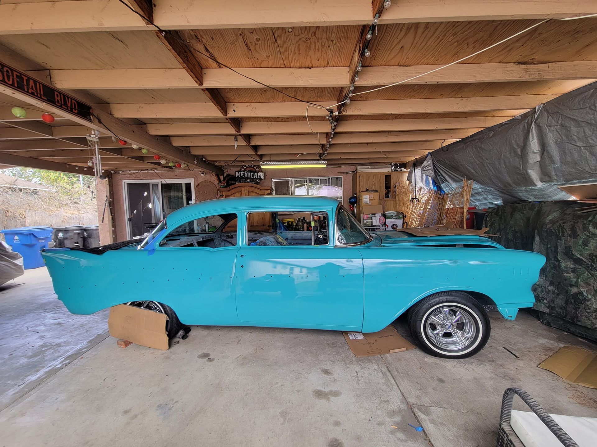 Blue classic car being restored under a wooden carport.