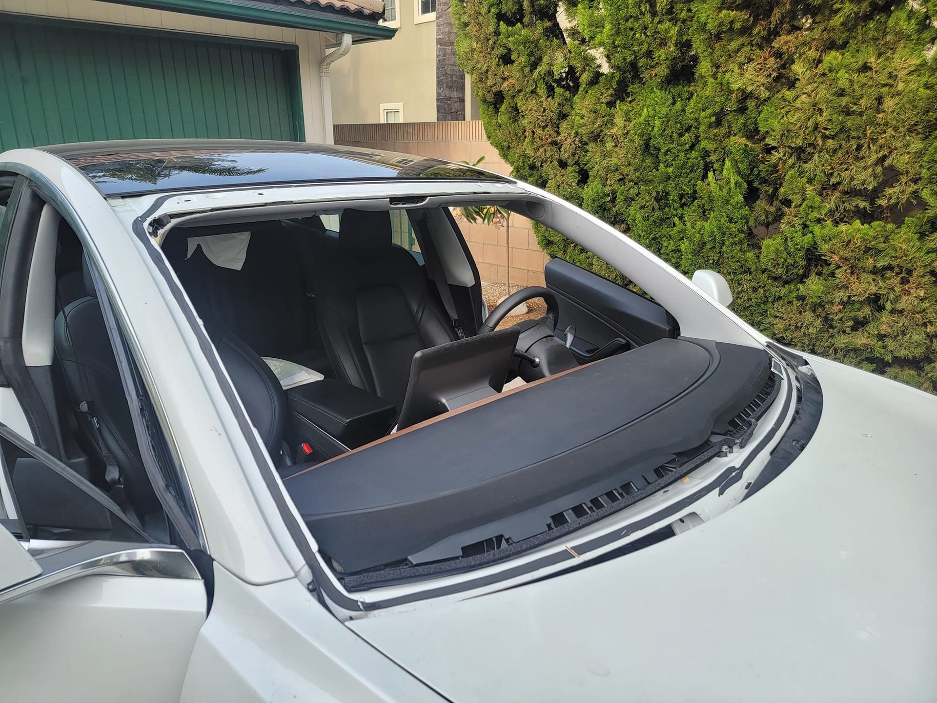 White car with a shattered windshield, parked outside a house with green foliage.