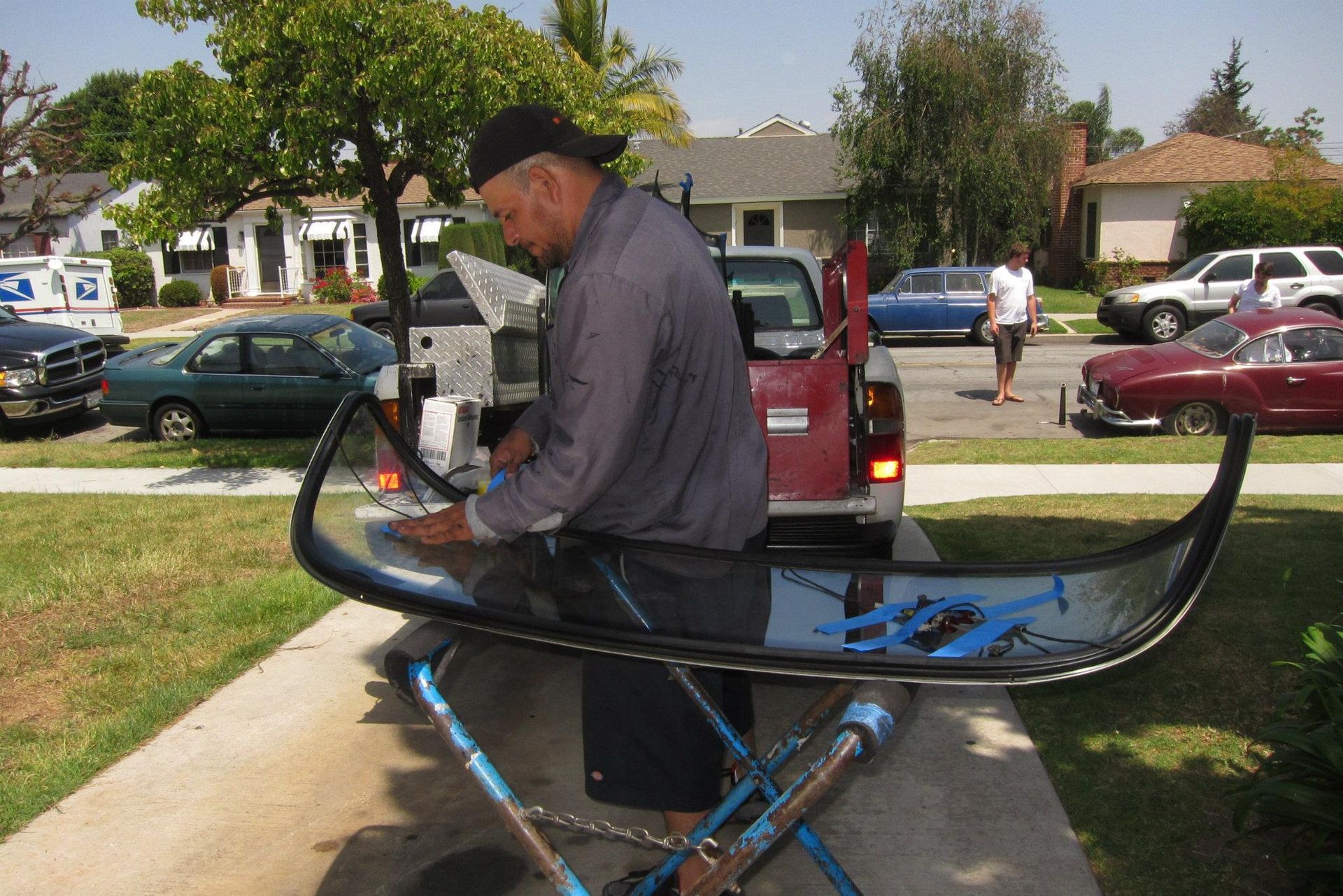 Man working on a car windshield supported by a stand, in front of a pickup truck, on a driveway.