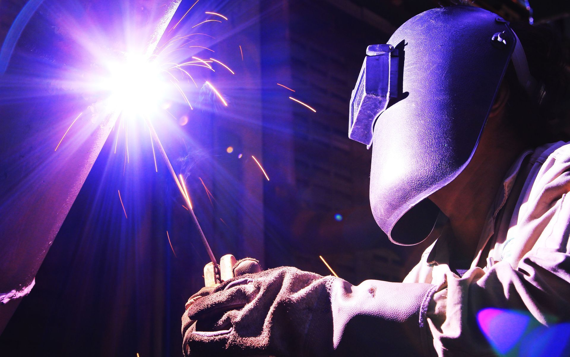 Welder in protective mask and gloves, using welding torch, bright light and sparks.