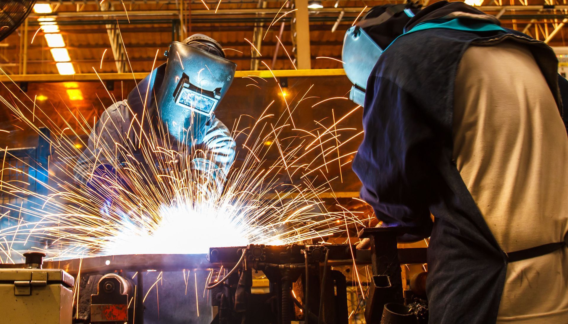 Two welders in protective gear, sparks flying as they work on metal in a factory.