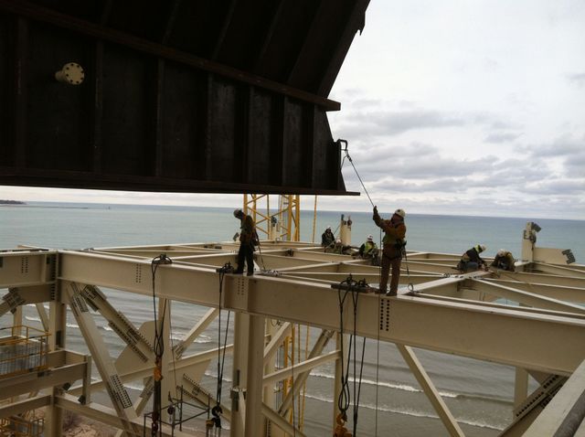 Workers installing a large metal beam on a construction frame near a body of water under a cloudy sky.