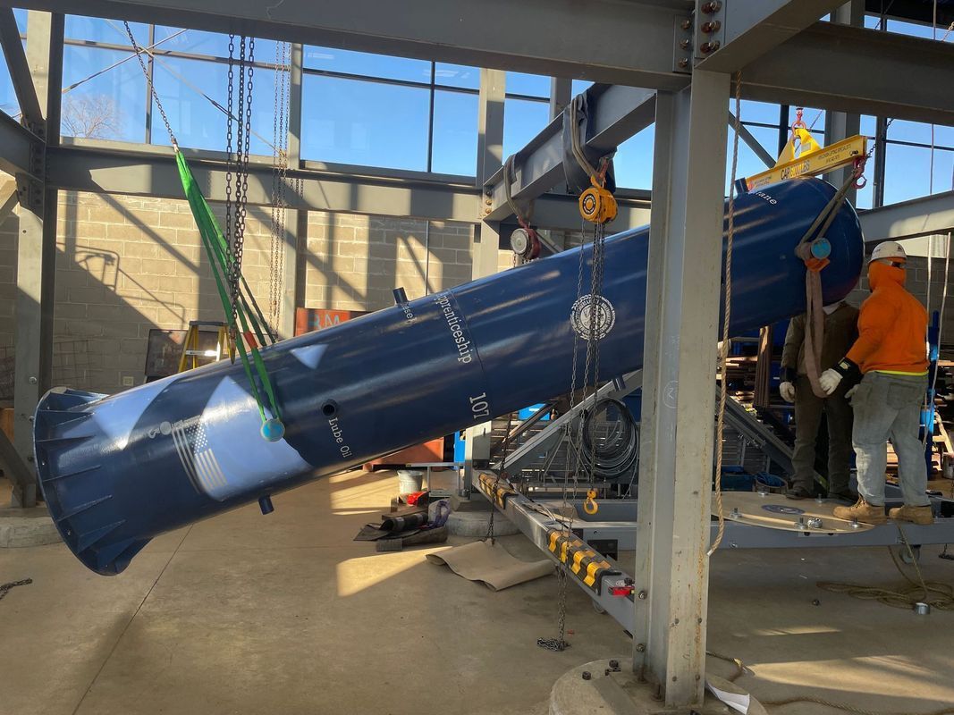 A large blue metal cylinder being lifted by a crane on a construction site; a worker in orange jacket nearby.