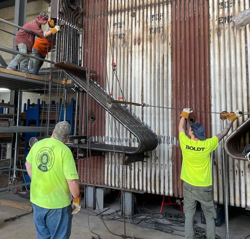 Workers installing industrial boiler tubes. Men in safety gear near large metal structure.