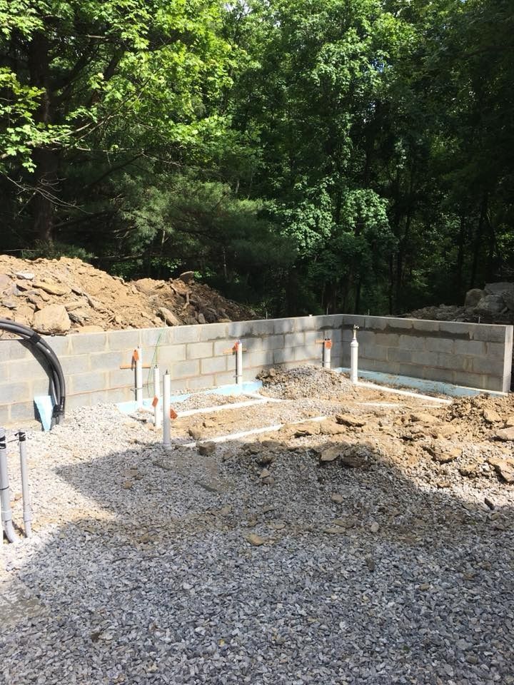 Construction site with cinder block walls, exposed plumbing, and gravel ground. Trees in the background.