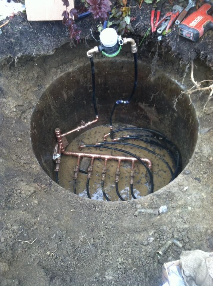 Open dirt pit containing copper and black irrigation piping, a water meter, and tools.
