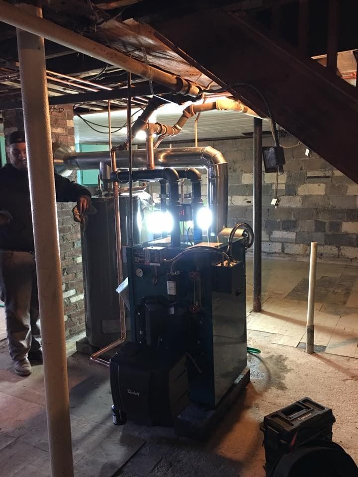 Man points at furnace in a basement setting. Pipes, water heater, and cinder block walls visible.