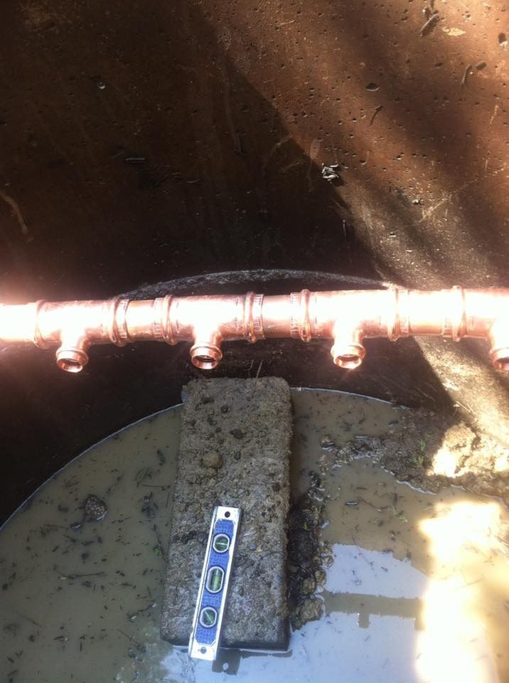 Copper pipes with T-fittings, likely part of a plumbing system, are being installed in a muddy trench.