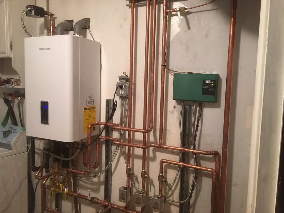White wall-mounted water heater with copper pipes in a utility room. Green control box is nearby.