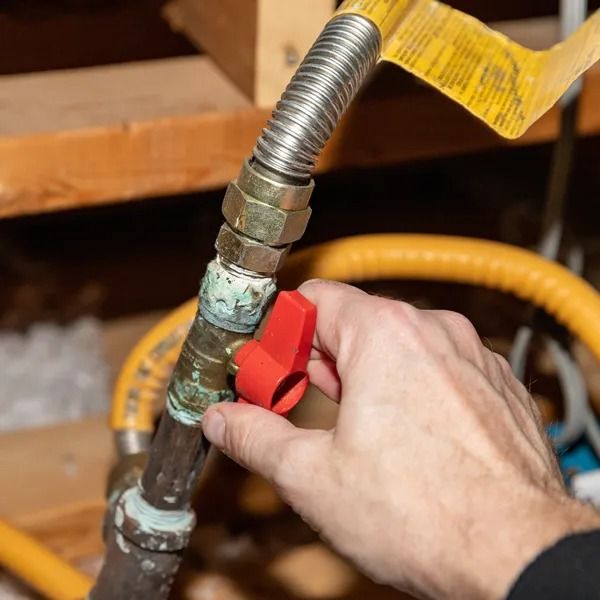 Hand turning a red gas valve, connected to a metal gas line, in a utility room setting.