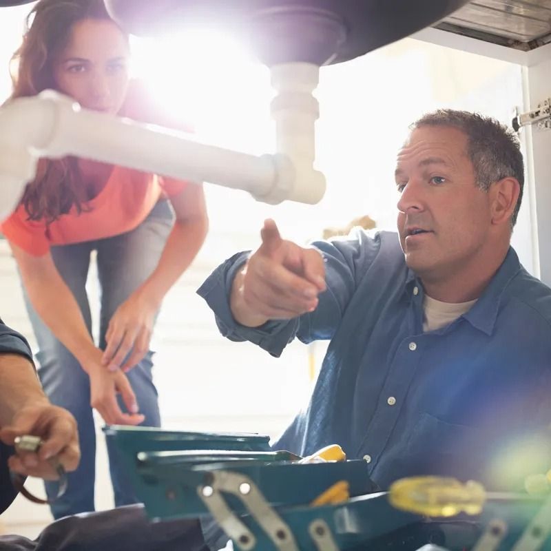 Man pointing at pipes under a sink; woman watches, toolbox visible.