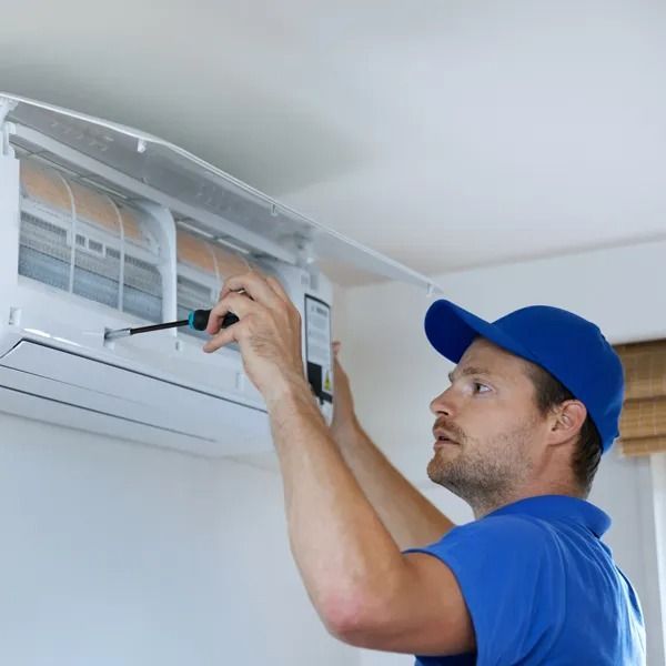 Man in blue uniform fixing a wall-mounted air conditioning unit with a screwdriver indoors.