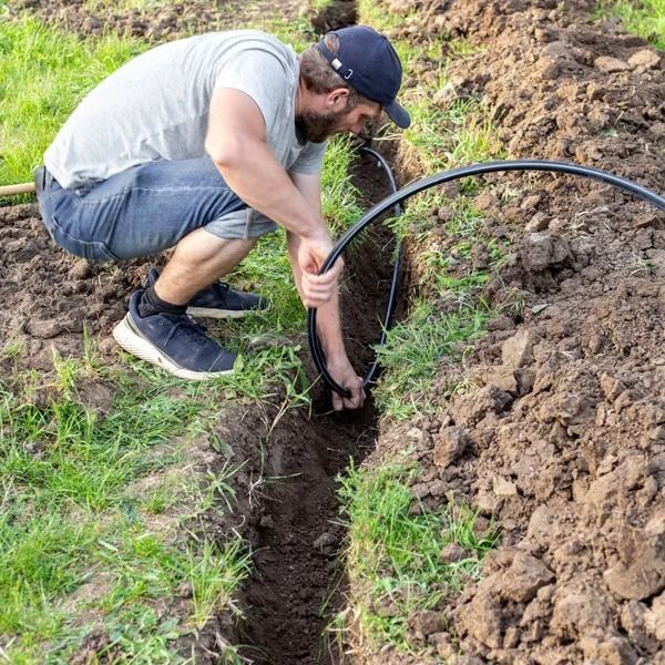 Man installing black irrigation tubing in a narrow garden trench outdoors.