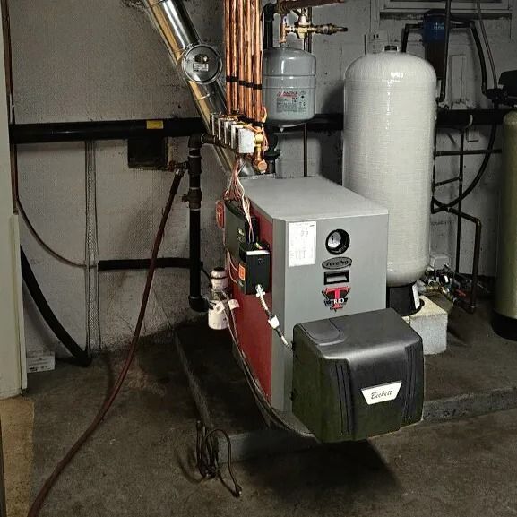 Boiler system in a basement. Grey and black machine with copper pipes, a white tank, and black wiring.