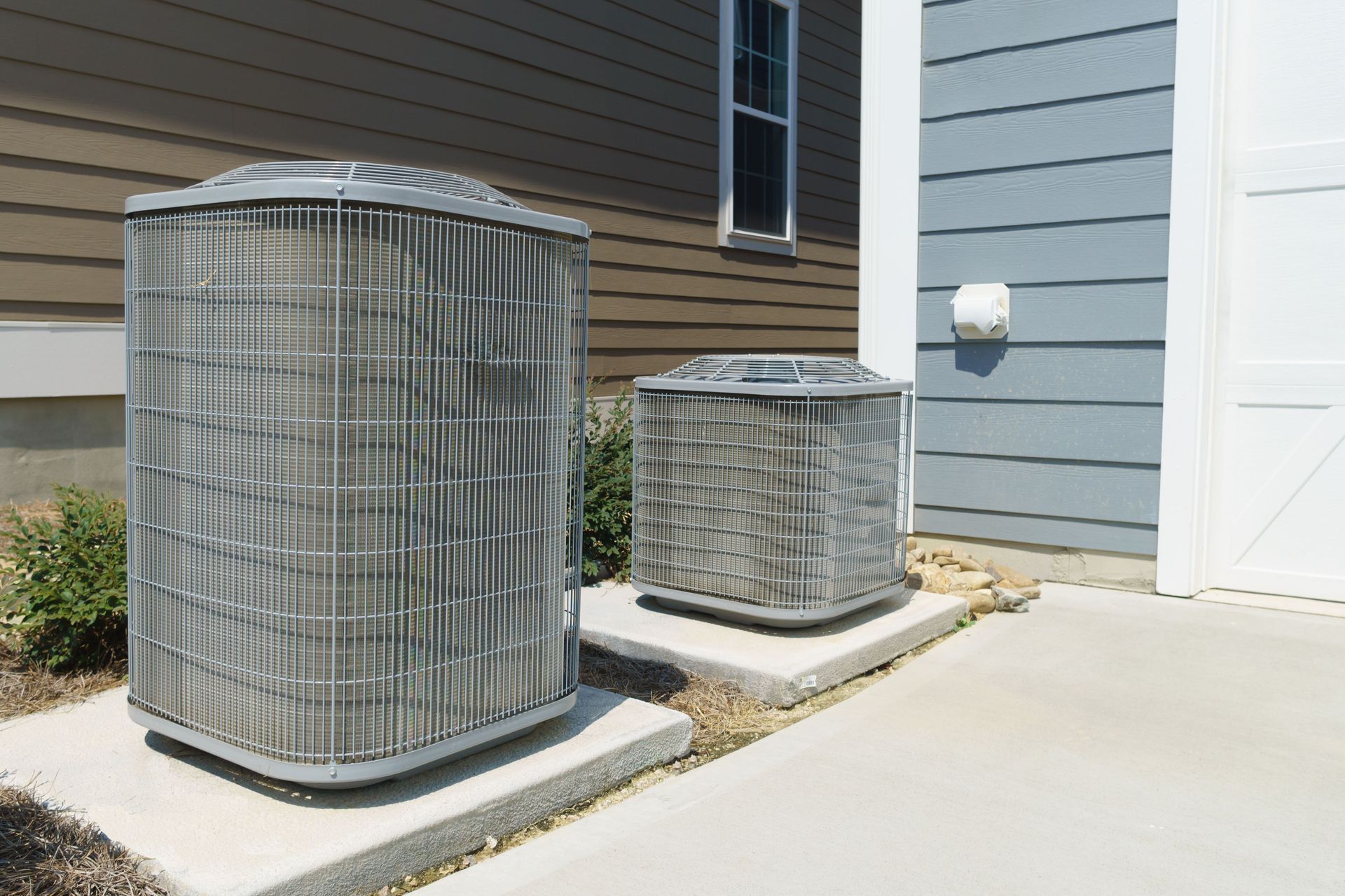 Two HVAC outdoor condenser units of different heights sitting on concrete pads next to a house exterior.