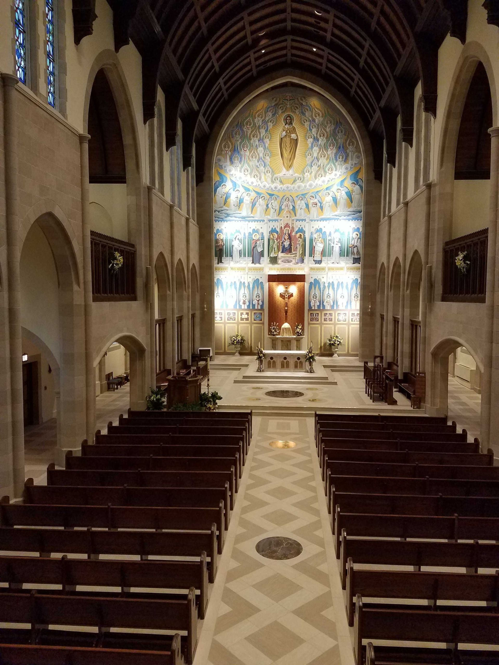 The inside of a church with rows of wooden benches and a mosaic on the wall.