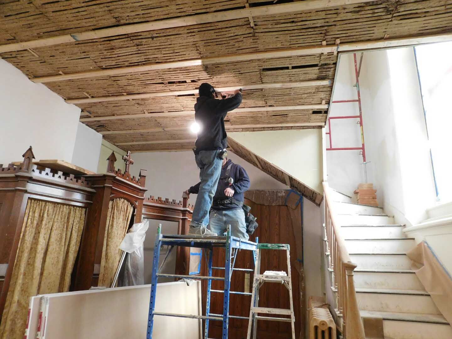 Two men are working on a wooden ceiling in a room with stairs.