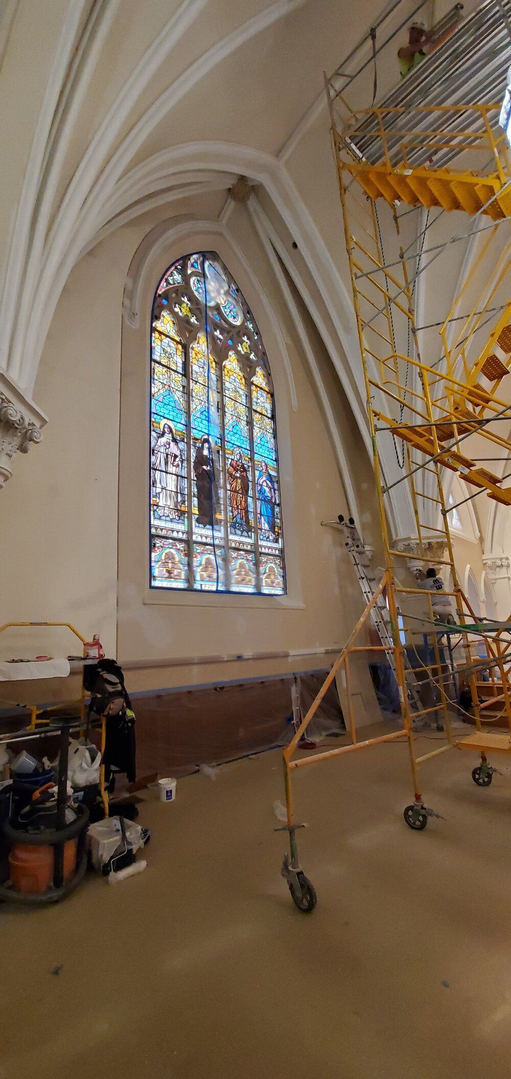 A stained glass window in a church with a ladder in the background.