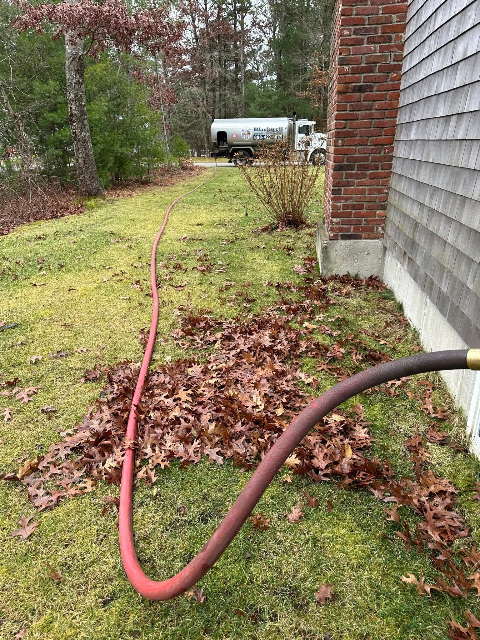 Red hose across a grassy yard beside a brick wall, leading to a white tank in the distance.