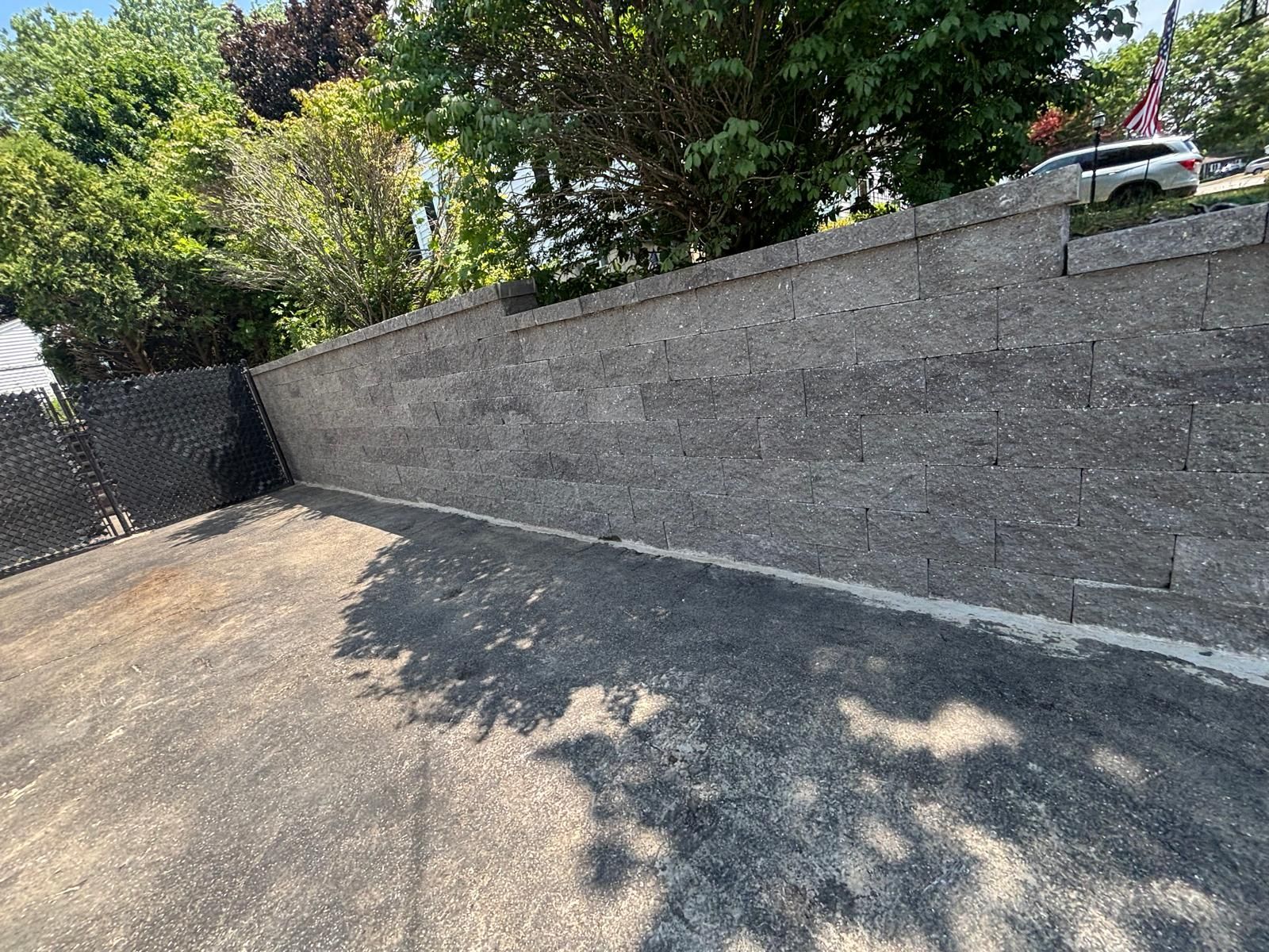 Empty paved driveway beside a gray retaining wall, with trees and sunlight shadows overhead