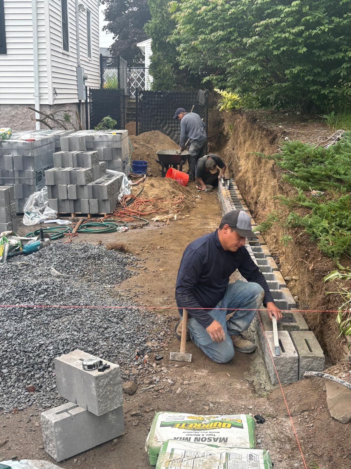 Construction workers building a retaining wall with concrete blocks outdoors. One is measuring, others are working.