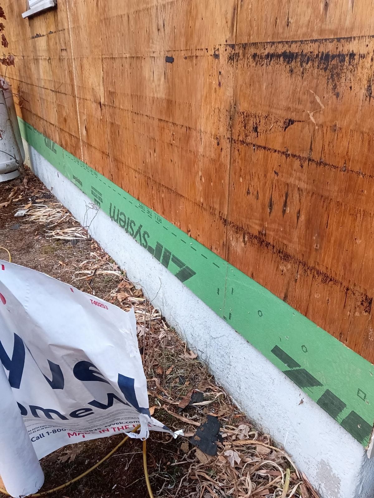Exterior wall construction with wood sheathing, green flashing, and concrete foundation. Debris is on the ground.
