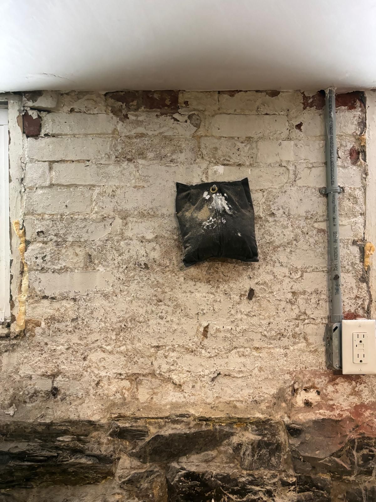 A dark bag attached to a white brick wall in a basement. Electrical conduit and outlet present.