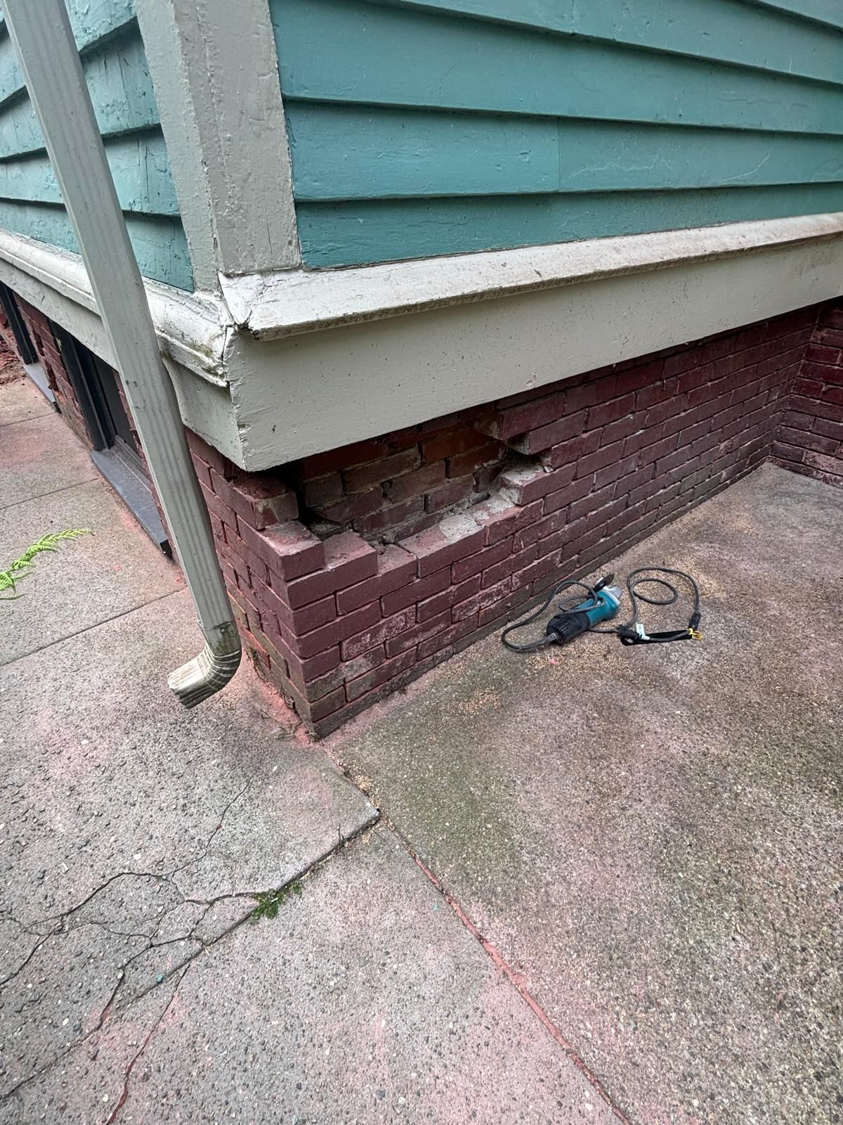 Damaged brick foundation beneath a teal house with a gutter, and a sidewalk.