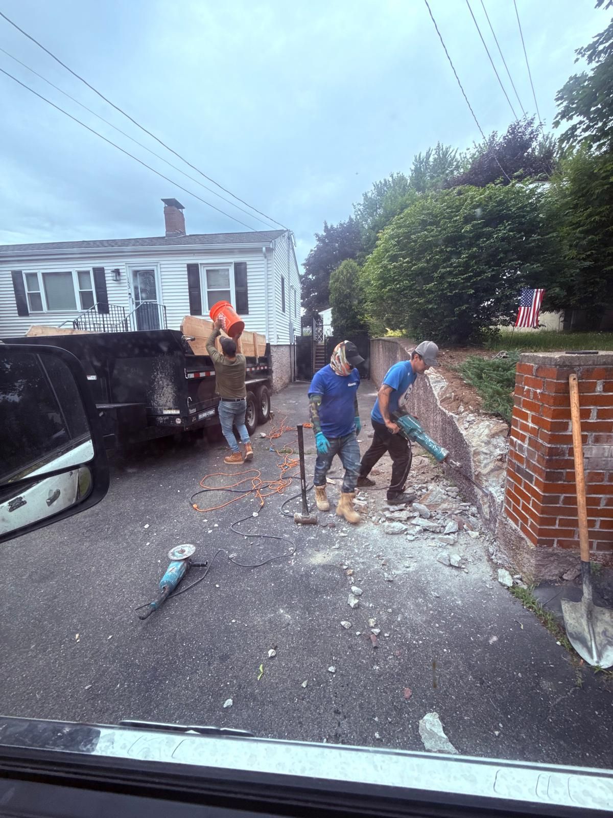 Construction workers demolishing a brick wall with tools; one carries items from truck on a driveway.