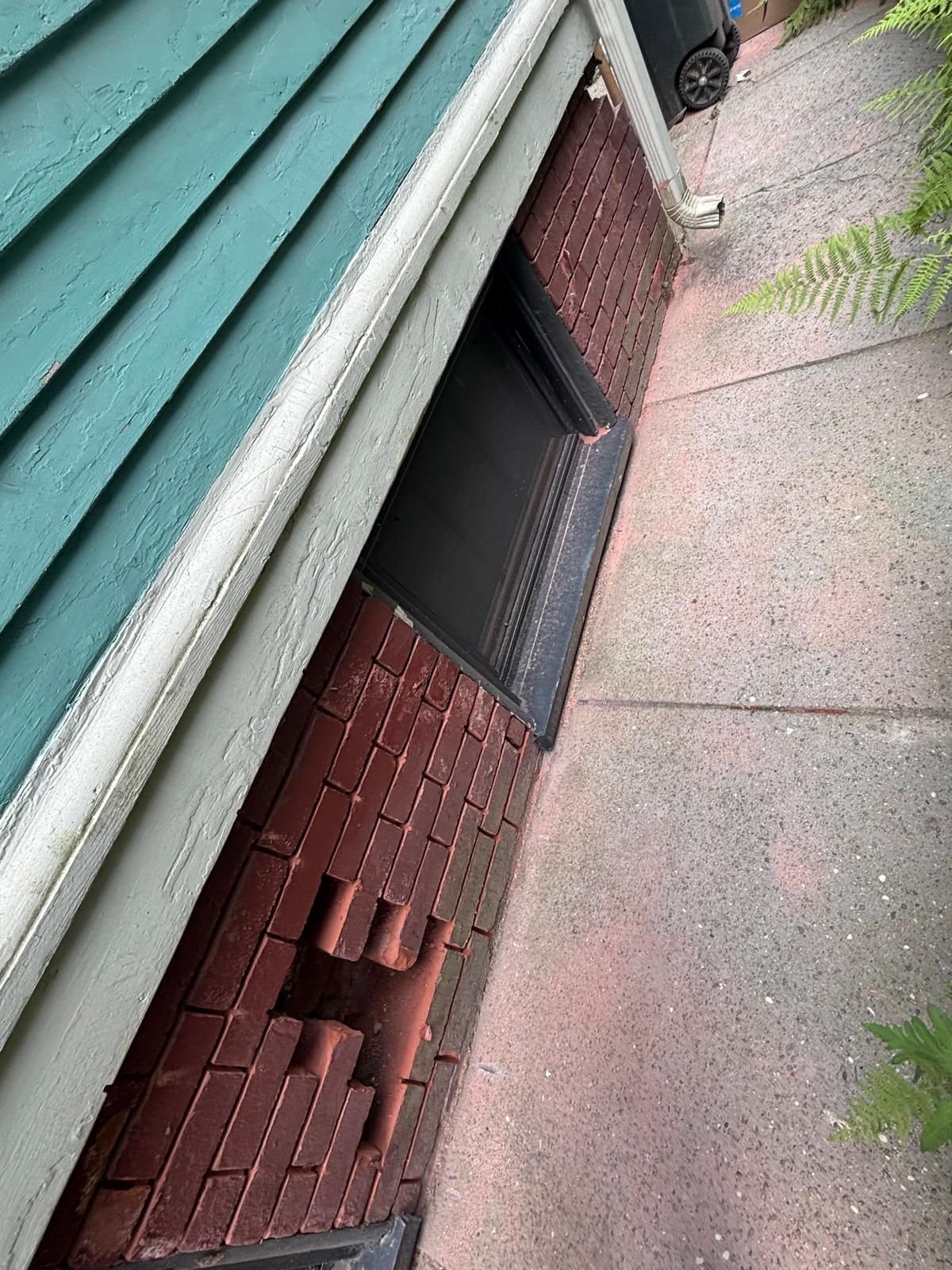 Brick wall with a small window under a turquoise roof, next to a concrete path.