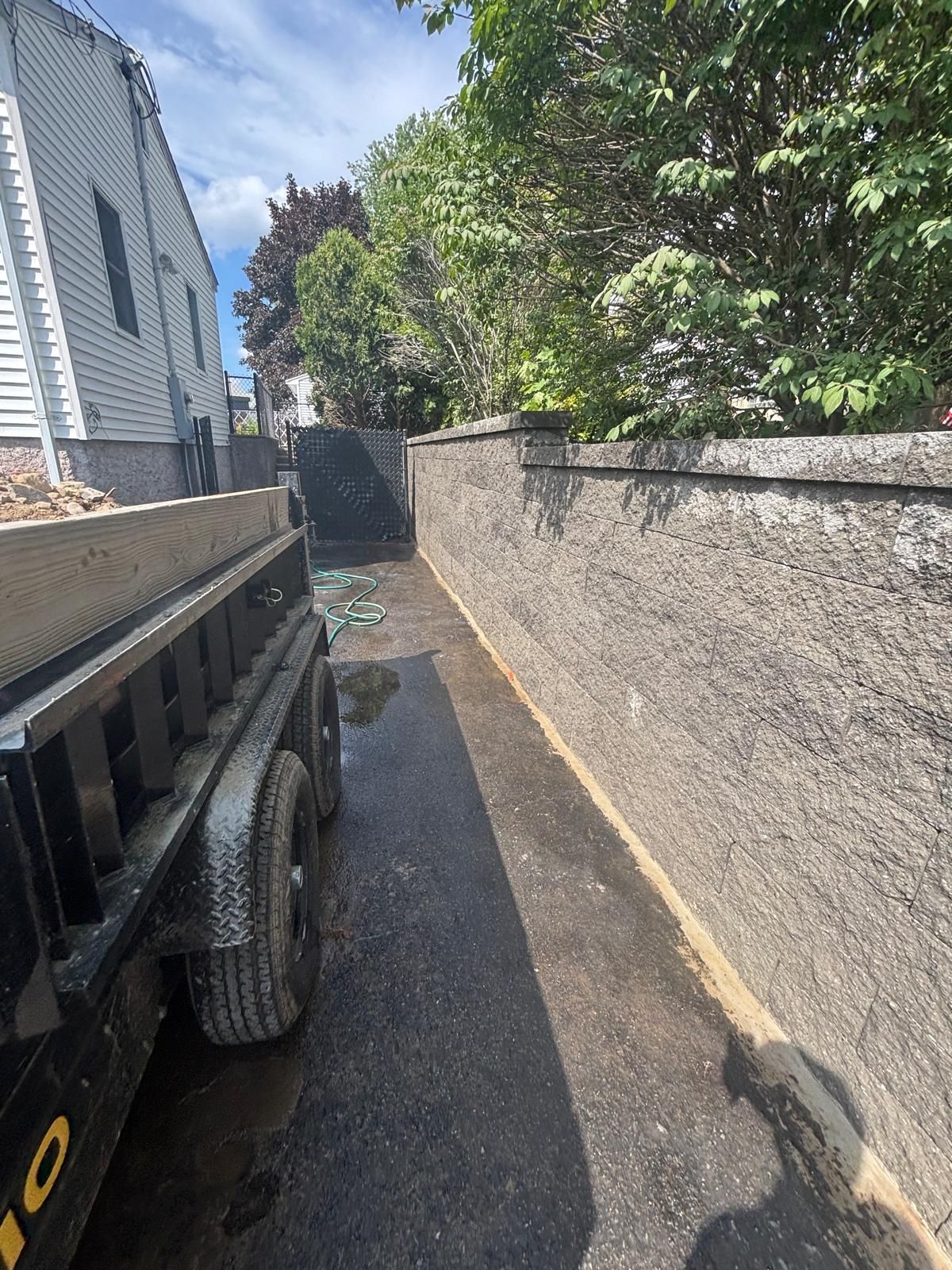 A black trailer on asphalt next to a textured concrete wall, trees, and a light-colored house.