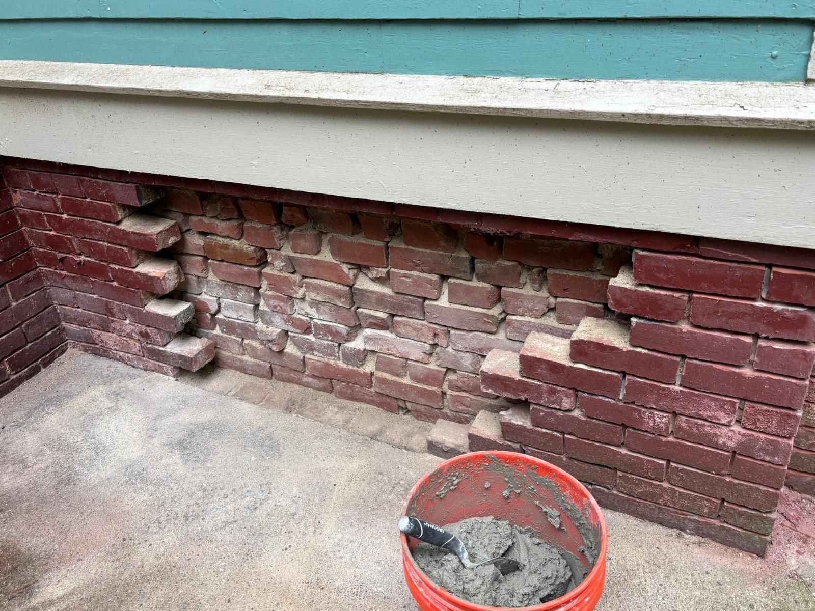 Damaged brick foundation being repaired; mortar in orange bucket.