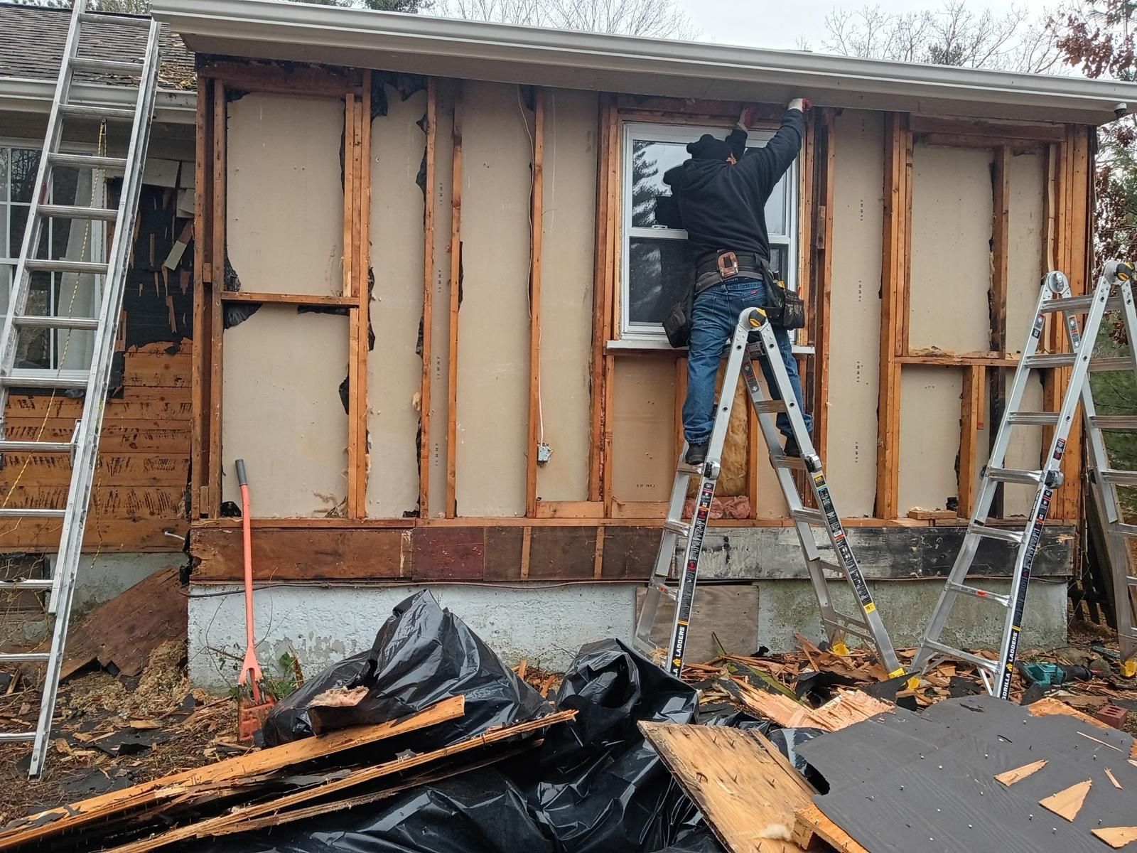 Person on ladder working on house exterior with exposed wall studs. Debris, ladders, and window visible.
