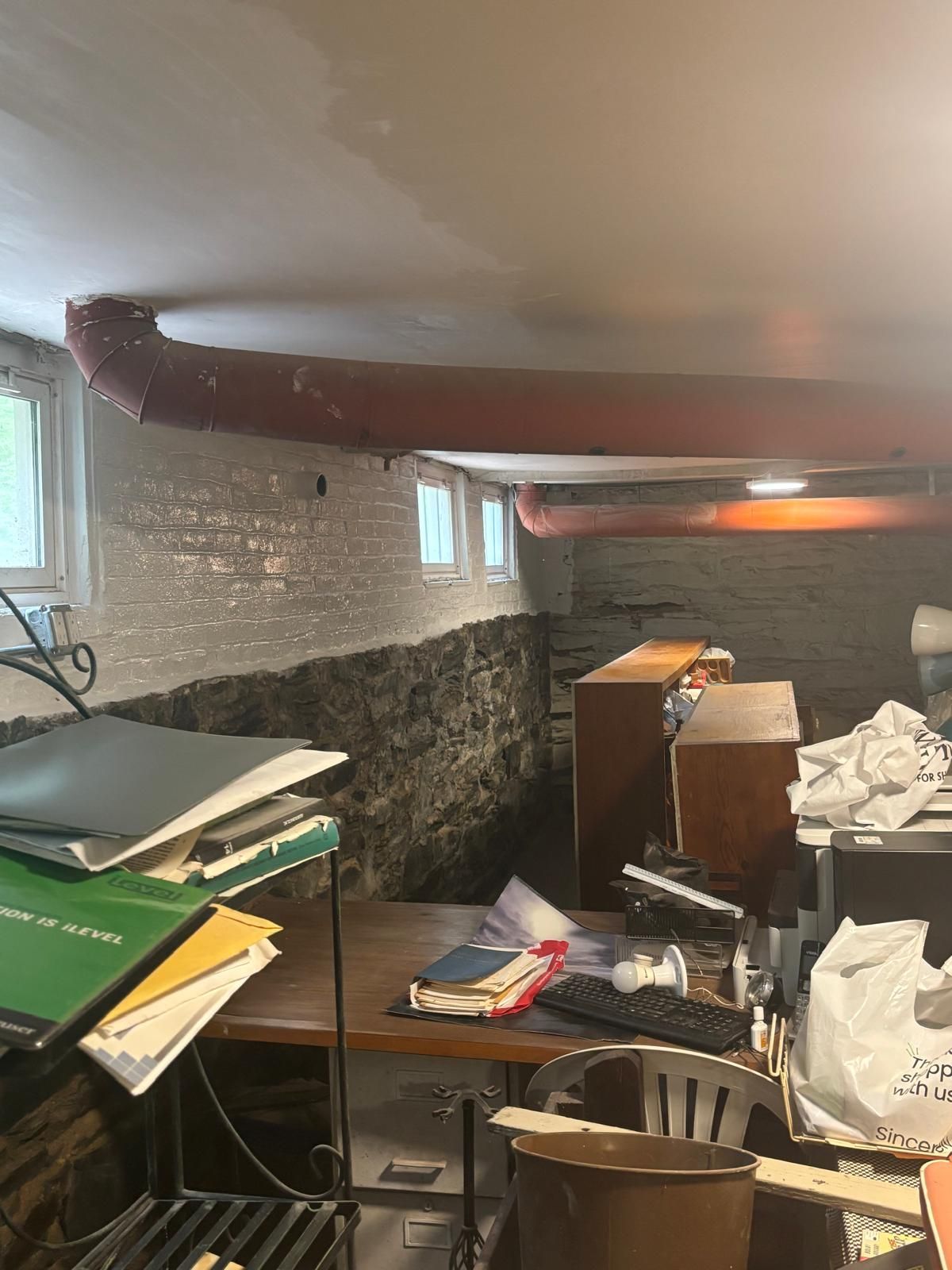 Cluttered basement with exposed brick wall, red ductwork, and small windows; desks and objects create visual clutter.