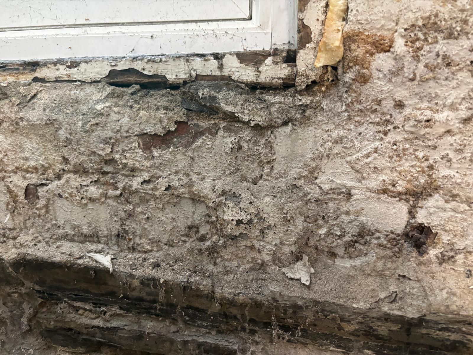 Damaged exterior brick wall beneath a window, with flaking paint and crumbling mortar.