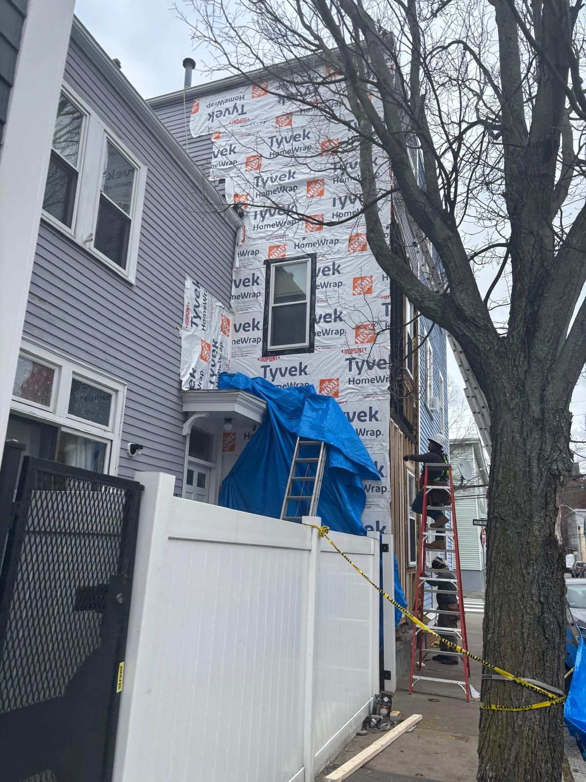 Building side undergoing construction. Tyvek wrap, blue tarp, ladder, and white fence are visible.
