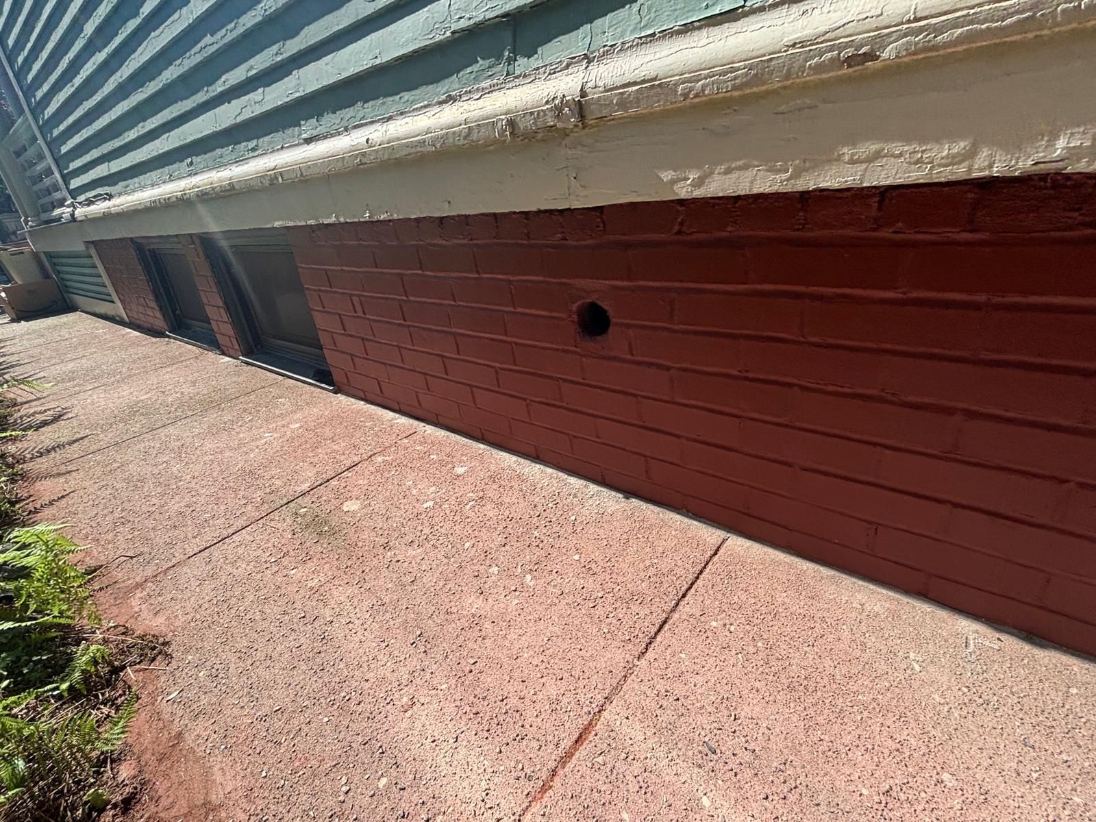 Red brick foundation with a black vent hole below a blue house with white trim, on a concrete path.