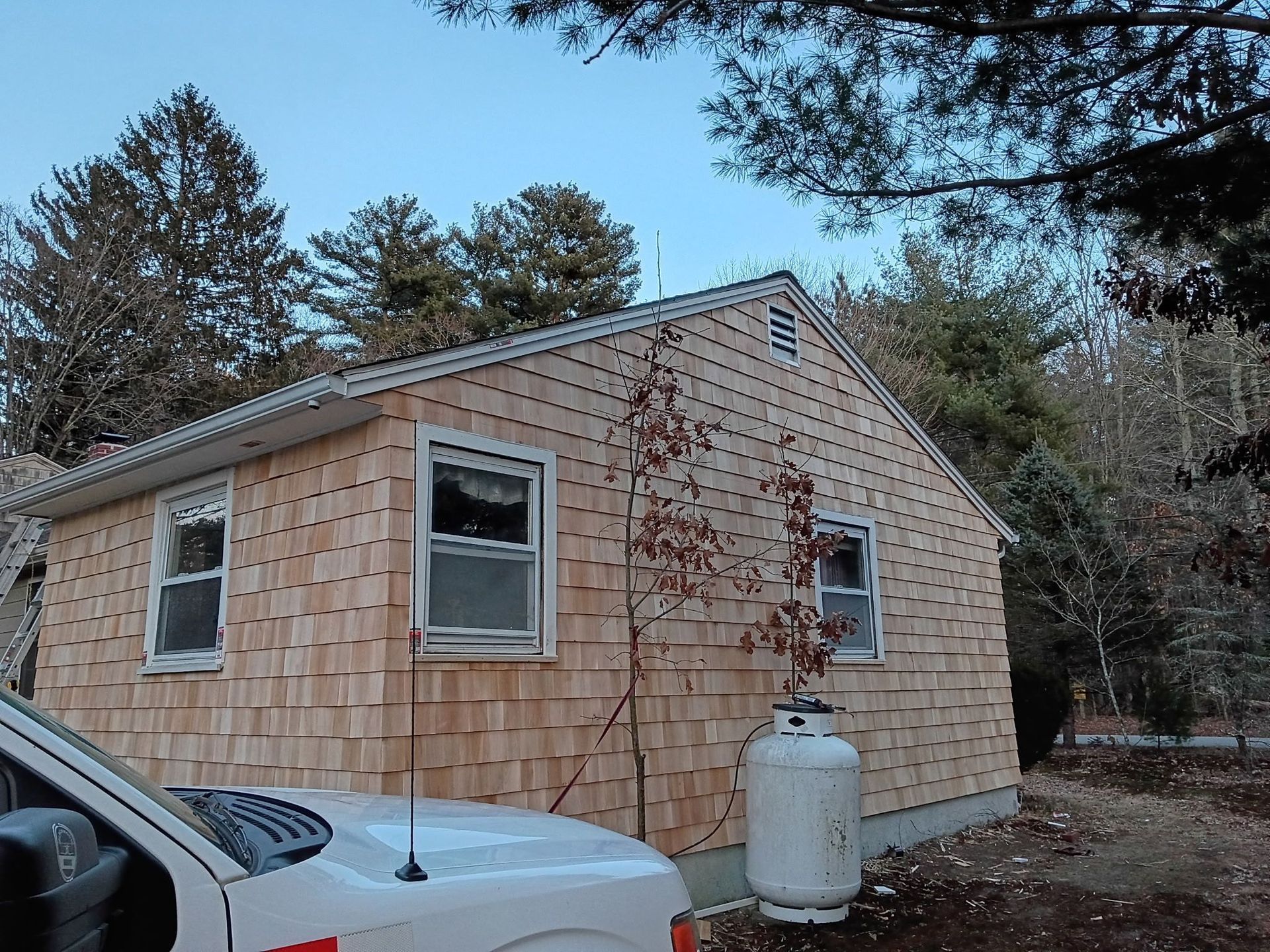 Small, wood-shingled building with two windows, a propane tank, and a white truck in front.