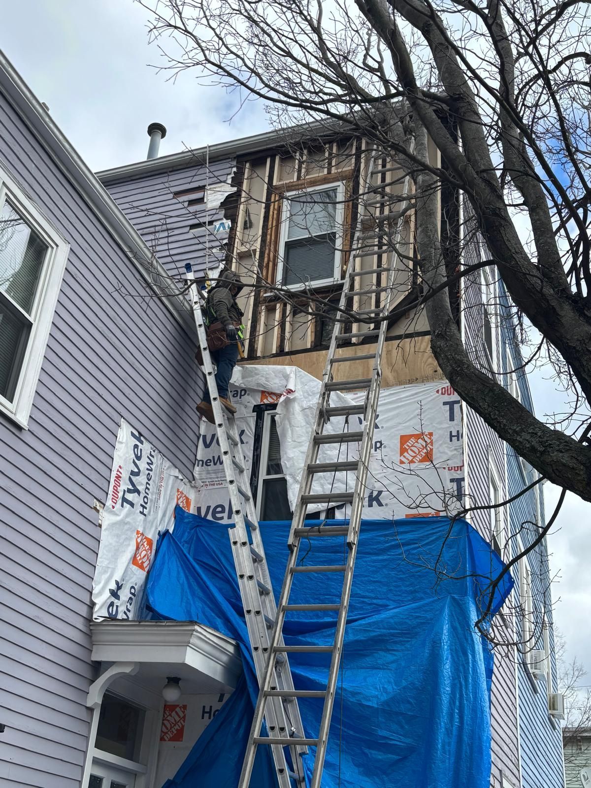 Workers on ladders repairing storm damage on a house exterior, covered with blue tarp and white wrap