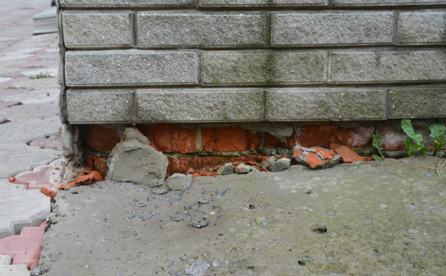 Damaged brick wall with crumbling mortar and a cracked concrete sidewalk below