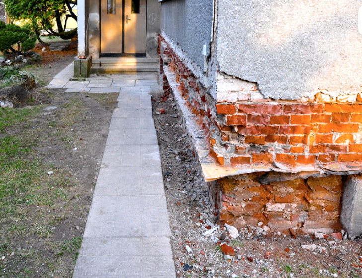 Cracked exterior wall with exposed red bricks beside a narrow concrete walkway to a doorway