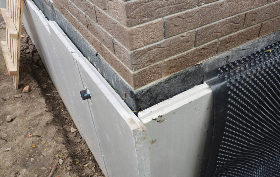 Damaged building corner with white siding, black waterproofing wrap, and exposed brick above soil and debris